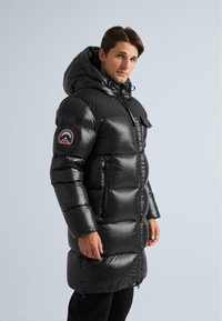 Muž oblečený v dlhom čiernom pufferskom kabáte s kapucňou a horskou témou na rukáve, hľadí smerom na kameru.