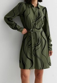Vestido de camisa verde oliva hecho de una tela suave, con mangas largas, botones en la parte delantera, un cinturón en la cintura y patrones abstractos en negro con forma de olas.