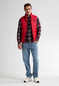 Rode bodywarmer over een geruit overhemd, blauwe spijkerbroek en witte sneakers. Heeft een doorgestikte textuur en een rits. Moderne, casual gelaagde look.