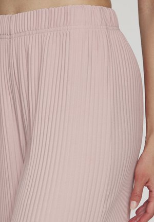 Nahaufnahme einer Person, die hellrosa, gerippte Strickhosen mit elastischem Bund trägt, wobei ein Teil ihrer Hand und Taille zu sehen ist.