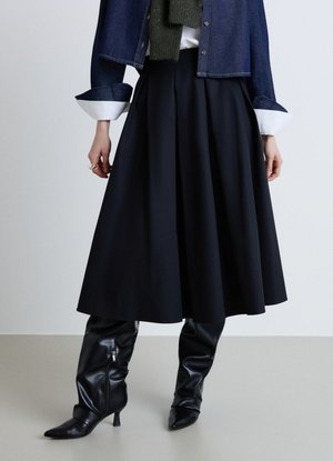 Persona che indossa una gonna midi nera, stivali neri alti fino al ginocchio con tacco, giacca in denim scuro e sciarpa verde scuro, in piedi contro uno sfondo neutro.