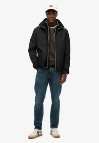 Czarna kurtka z zapięciem na zamek i kapturem, szary sweter pod spodem, niebieskie dżinsy i białe trampki z czarnymi akcentami. Kremowa czapka.