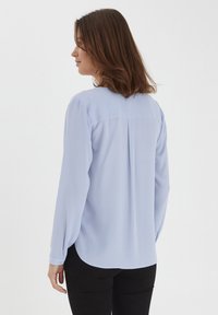 Camicia a maniche lunghe di un blu chiaro, con una texture liscia, scollo rotondo e una sottile piega sul retro. Indossata con pantaloni neri aderenti.