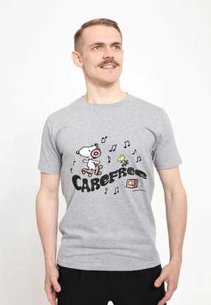 Młody mężczyzna z wąsami, ubrany w szary t-shirt z wizerunkami Snoopy'ego i Woodstoka, jeżdżący na wrotkach z nutami muzycznymi i napisem "CAREFREE."