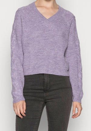 Femme portant un pull en maille violet clair en V avec des motifs torsadés sur les manches et un pantalon noir taille haute sur fond blanc.