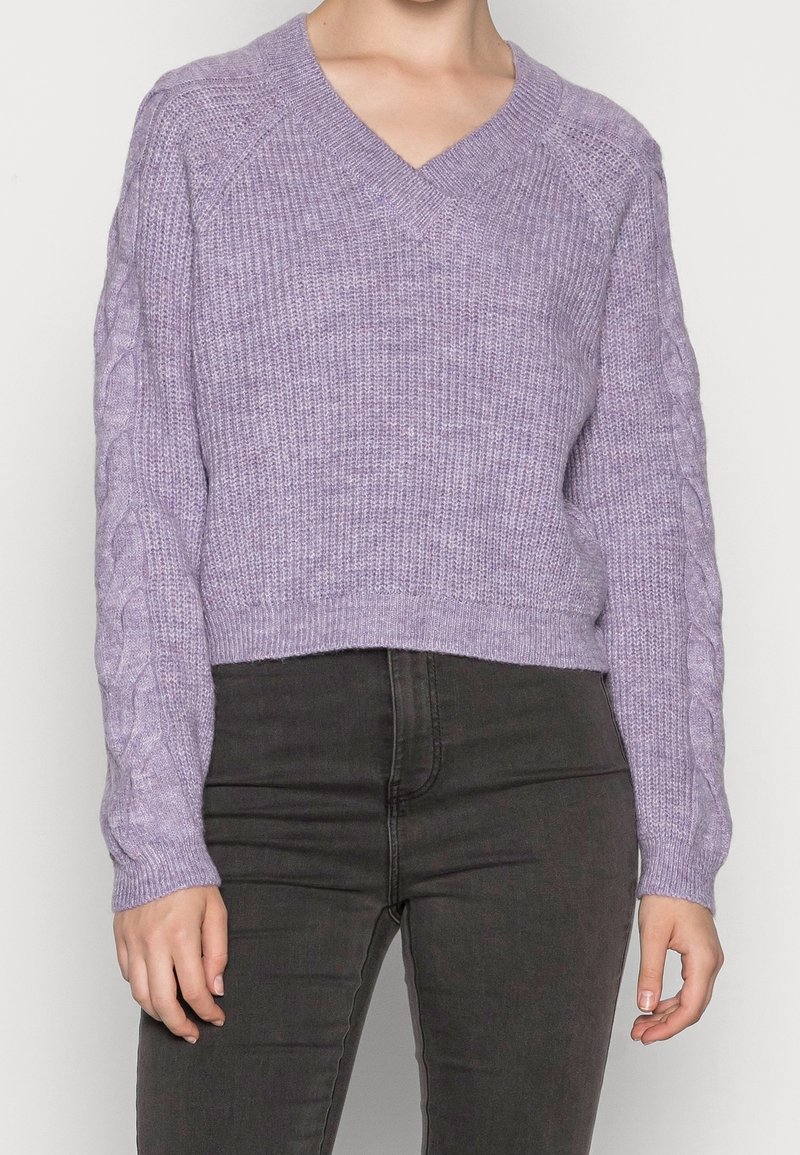 Femme portant un pull en maille violet clair en V avec des motifs torsadés sur les manches et un pantalon noir taille haute sur fond blanc.