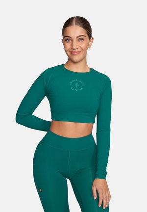 Frau lächelt, trägt ein türkisfarbenes, langärmliges Crop-Top und passende, hoch taillierte Leggings mit dem "Gold's Gym"-Logo oben darauf.