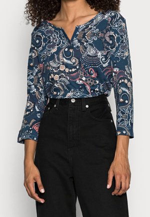 Frau trägt ein marineblaues, langärmliges Oberteil mit floralem Paisley-Muster, in hoch taillierte schwarze Jeans gesteckt, steht mit entspannten Armen.