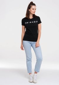 Black cotton T-shirt featuring white "F.R.I.E.N.D.S" text across the chest. Worn with light blue jeans and white sneakers.