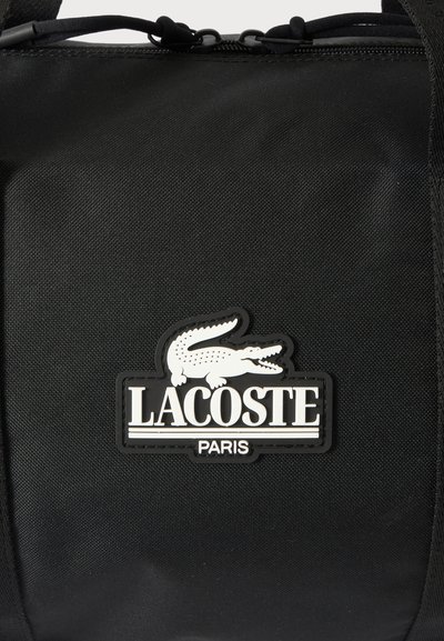 Gros plan sur un sac noir texturé avec un écusson blanc représentant le crocodile Lacoste et le logo Paris.