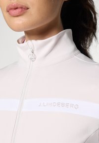 Lyserød zip-up jakke med høj krave, prydet med en hvid stribe og "J. Lindeberg" logo broderet i matchende farve på brystet.