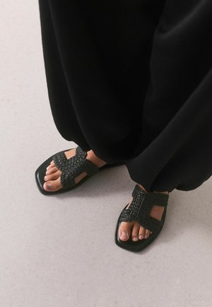 Fødder iført sorte vævede læder sandaler og løse sorte bukser på en lysegrå gulv.