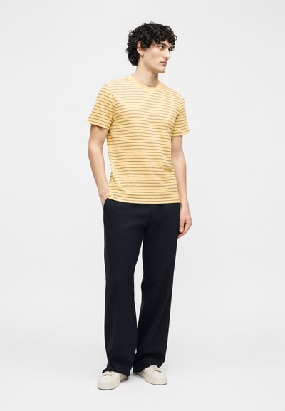 Άνδρας που στέκεται με τα χέρια στις τσέπες, φορώντας κίτρινο ριγέ μπλουζάκι, μαύρο παντελόνι και λευκά αθλητικά παπούτσια, σε απλό φωτεινό φόντο.