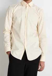 Camicia button-up gialla chiara con colletto, maniche lunghe e orlo leggermente curvo. Tessuto morbido con una texture liscia.