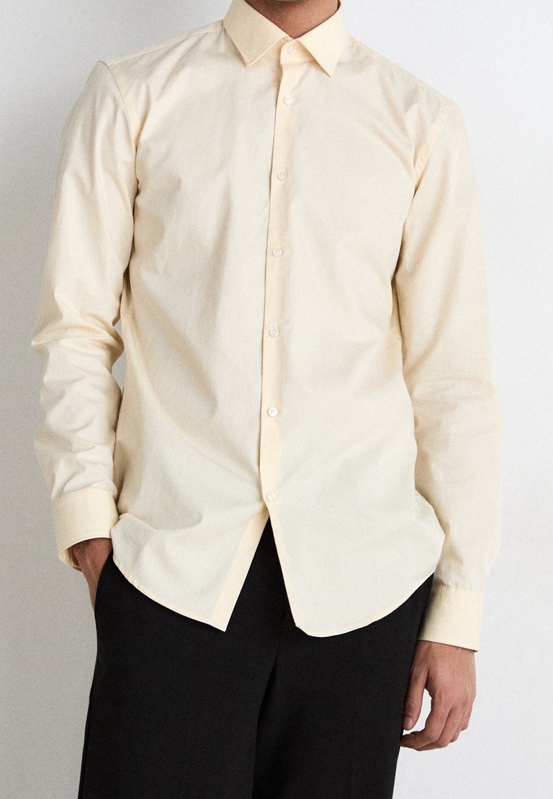 Camicia button-up gialla chiara con colletto, maniche lunghe e orlo leggermente curvo. Tessuto morbido con una texture liscia.