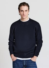Marineblauer Pullover mit strukturierter Strickoptik, Rundhalsausschnitt und gerippten Bündchen. Wird über einem weißen Hemd getragen und mit blauen Jeans kombiniert.