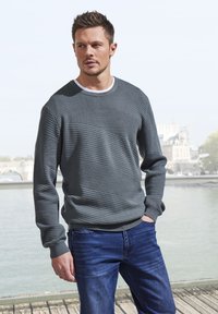 Grauer, strukturierten Pullover mit horizontalen Streifen, Rundhalsausschnitt und langen Ärmeln. Kombiniert mit blauen Jeans, steht nahe einem Gewässer.