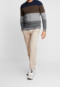 Neulepusero, jossa on tummansinisiä, ruskeita, harmaita ja vaaleanharmaita vaakasuoria raidoituksia, yhdistettynä béžheissä housuissa ja valkoisissa lenkkareissa.