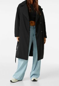 Svart lång kappa med en avslappnad passform, stor krage och tryckknappsstängning, kombinerad med ljusblå vida jeans och krämfärgade sneakers.