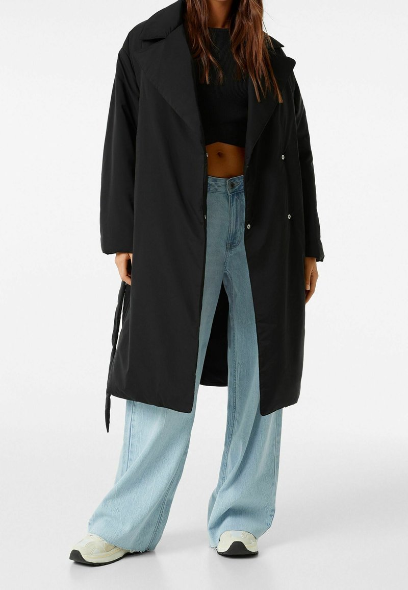 Svart lång kappa med en avslappnad passform, stor krage och tryckknappsstängning, kombinerad med ljusblå vida jeans och krämfärgade sneakers.