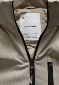 Veste imperméable beige avec une texture subtile, zips noirs et une étiquette indiquant la marque "Jack & Jones" et des caractéristiques telles que la résistance à l'eau.