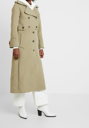 Persona vistiendo un abrigo largo beige de doble botonadura con botones, pantalones blancos de pierna ancha y botines blancos con tacón sobre un fondo liso.