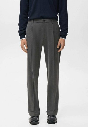 Man wearing dark gray tailored trousers with cuffs, black shoes, and a navy blue long-sleeve top, standing against a plain background.