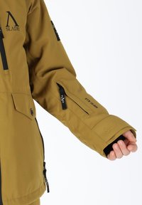 Braune wasserdichte Jacke mit einer Reißverschlusstasche am Ärmel. Verfügt über ein Logo und verstellbare Manschetten. Das Material wirkt glatt und langlebig.