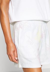 Detailní záběr ruky osoby vedle bílých šortek s jemným pastelovým vírovým vzorem a bočním kapsou, které jsou doplněny bílým topem.