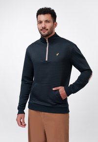 Quarter-zip hette i mørk marineblå med strukturmønster, beige glidelås og gullfarget fuglelogo. Har frontlommer og ribbestrikkede mansjetter.