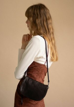 Borsa a mano nera in pelle con forma curva, tracolla regolabile e hardware argento. Il modello indossa una maglia bianca a maniche lunghe e un vestito floreale marrone.
