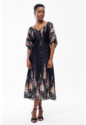 Kobieta w czarnej sukience midi z guzikami z przodu i różowymi kwiatowymi wzorami, do tego czarne szpilki z paskiem wokół kostki oraz wiszące kolczyki.