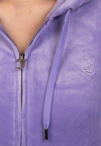 Purple zip-up hoodie featuring a soft, textured fabric, silver zipper with a decorative pull, and a small embroidered logo on the chest.