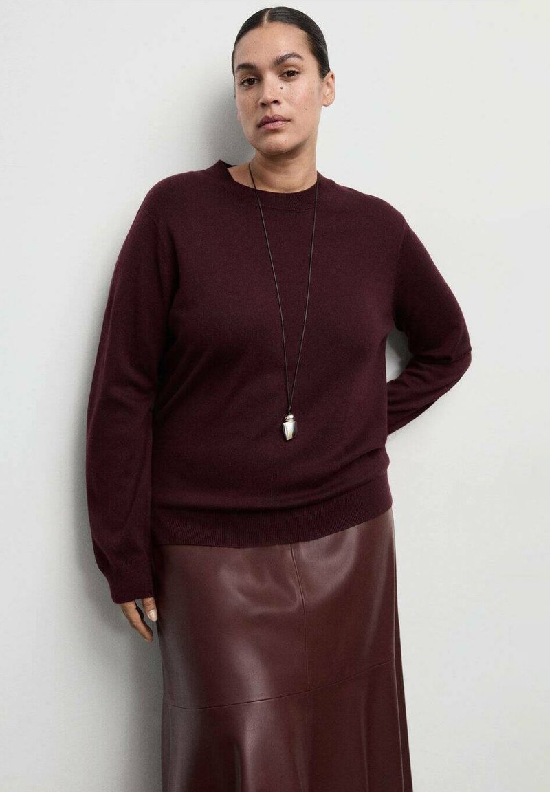 Pull en tricot bordeaux à col rond, associé à une jupe en cuir marron. La tenue est agrémentée d'un long collier pendentif argenté.