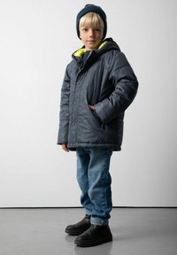 Młody chłopiec w ciemnym czapce, szarej zimowej kurtce z kapturem i żółtym podszyciem, niebieskich dżinsach i czarnych butach stoi na szarym podłodze w pobliżu białej ściany.