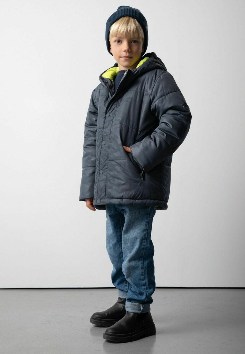 Młody chłopiec w ciemnym czapce, szarej zimowej kurtce z kapturem i żółtym podszyciem, niebieskich dżinsach i czarnych butach stoi na szarym podłodze w pobliżu białej ściany.