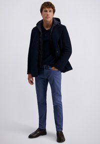Marineblauer, strukturiert aussehender Peacoat über einem dunklen Polo, kombiniert mit hellblauen Chinos und dunkelbraunen Slippern, mit einer Kapuze und Knopfdetails.