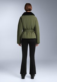 Olivno zelena podložena jakna s crnim umetnim krznom oko vrata, strukirana, s dugim rukavima, u kombinaciji s crnim širokim hlačama s patentnim zatvaračima.