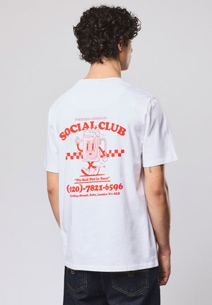 Person mit lockigem Haar trägt weißes T-Shirt mit rotem "SOCIAL CLUB"-Schriftzug, Cartoon-Bierkrug und Kontaktdaten aus London auf der Rückseite.