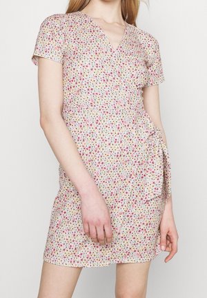 Femme portant une robe portefeuille à manches courtes et décolleté en V, avec une ceinture nouée sur le côté, ornée de petites fleurs roses, violettes et jaunes sur un fond clair.