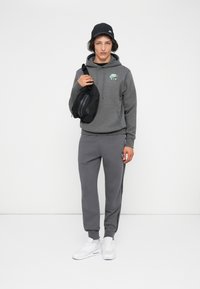 Jovem com um hoodie cinza da Nike e calças de moletom, tênis brancos, boné preto, segurando uma mala crossbody preta da Nike em frente a um fundo branco.