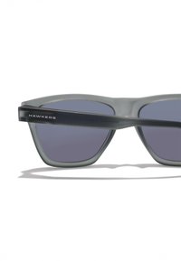 Szürke napszemüveg geometriai mintával, sötétlila lencsékkel és fekete szárakkal, amelyeken a "HAWKERS" márkanév látható.