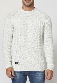 Krämfärgad stickad tröja med kabelmönster, rund halsringning och ribbade muddar och nederkant. Texturerad garn, strukturerad passform, avslappnad stil.