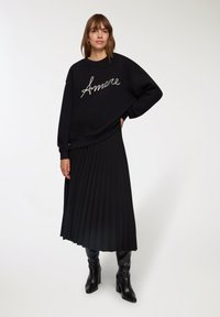 Czarny sweter z haftem "Amore" w kolorze białym, zestawiony z czarną plisowaną spódnicą midi i czarnymi botkami do kolan, na jasnym tle.