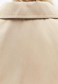 Detailní záběr béžového límce kabátu a horní části zad z textury látky s viditelnými švy.