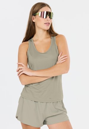 Ung kvinde med langt hår iført reflekterende store solbriller, ærmeløs beige top og matchende shorts, med armene krydset.