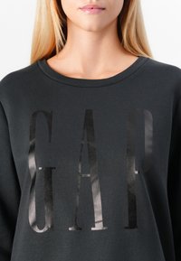 Dark gray sweatshirt featuring the word "GAP" in large, glossy black letters across the chest, with a round neck and soft texture.