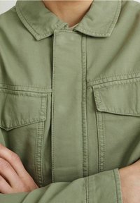 Olivově zelená bavlněná bunda s designem dvojitých kapes, knoflíkovým límcem a viditelnými detaily šití. Texturovaná látka, volný střih.