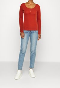 Röd långärmad topp med rundad halsringning och tre knappar, tillsammans med ljusblå skinny jeans och vita sneakers.