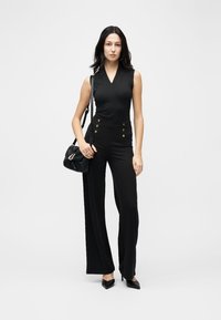 Femeie stând, purtând un top negru fără mâneci și pantaloni negri cu talie înaltă și picioare largi, cu nasturi aurii, ținând o geantă neagră cu bretea pe umăr.